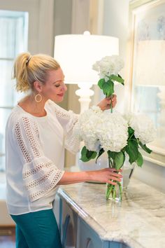 a woman arranging white flowers in a vase on a counter with a mirror behind her