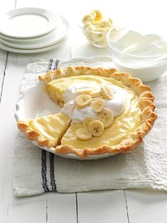 a pie with bananas and whipped cream on top sits on a white tablecloth next to plates