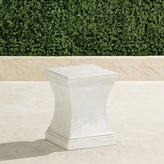 a white pedestal sitting on top of a cement floor next to a green hedge covered wall