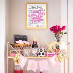 a bar cart filled with pink and gold items on top of a wooden table next to a framed movie poster