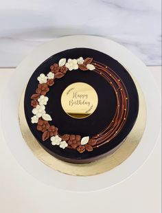 a black and gold birthday cake with white flowers on the top is sitting on a plate