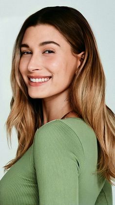 a woman with long hair smiling and wearing a green shirt, standing in front of a white wall