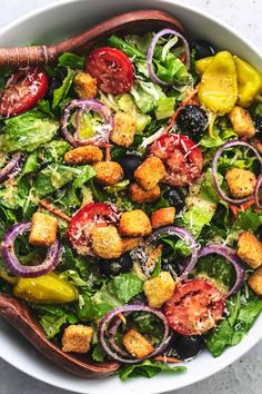 a salad with croutons, olives, tomatoes and other vegetables in a white bowl