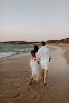 a man and woman walking on the beach holding each other's hands as they walk towards the water