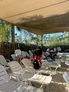 several people are sitting under an awning at the beach while others stand and sit on lawn chairs