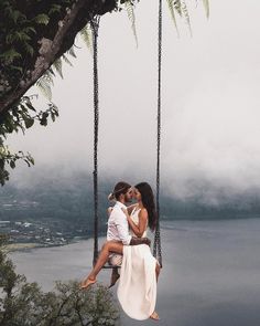 two people are sitting on a swing in front of the water and trees, one person is kissing the other