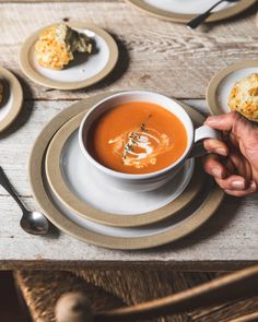 a person holding a cup of soup on top of a white plate with silverware