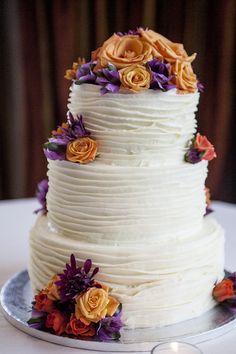 a three tiered white cake with orange and purple flowers on the top, next to a lit candle