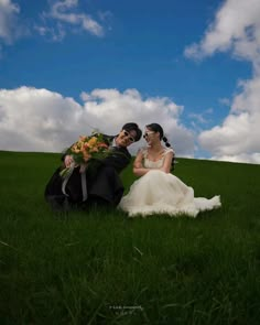 a bride and groom sitting in the grass under a blue sky with puffy clouds