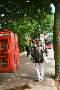 a woman is walking down the sidewalk next to two red telephone booths and some trees