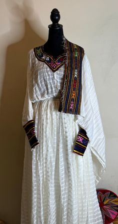 Ethiopians and Eritrean traditional dress (habesha kemis) dress is made of a white, textured fabric with intricate, colorful embroidery around the neckline, cuffs, and hem. The embroidery features geometric patterns in vibrant colors, which are typical of Ethiopian traditional attire. The dress is long, reaching the floor, and has a loose, flowing silhouette with an elasticized waist. It's a beautiful example of cultural fashion.any ceremony beautiful dress       Simple 100% comfortable dress Fashion Traditional Dresses, Ethiopian Fashion, Ethiopian Clothing, Cultural Fashion, Habesha Kemis, Fashion Traditional, Colorful Embroidery, Dress Simple, Ethnic Dress