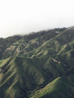 an airplane flying over the top of a green mountain covered in clouds and grass on a cloudy day