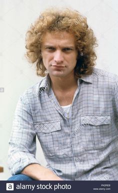 a man with curly hair sitting down and looking at the camera, in front of a white