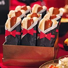 hot dogs wrapped in black paper with red bows are on a table next to other food