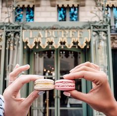 two people holding up small macaroons in front of a building with ornate doors