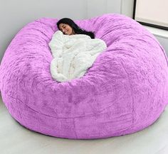 a woman wrapped in a blanket is sitting on a large purple bean bag chair that's next to a window