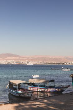 two boats are docked on the shore of a beach with mountains in the background and blue water