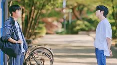 two young men standing next to each other on a sidewalk in front of trees and bicycles