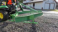 a green tractor is parked on gravel next to a house and barn, with the front end of it's loader attached