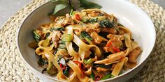 a white bowl filled with pasta and meat on top of a woven place mat next to a wooden spoon