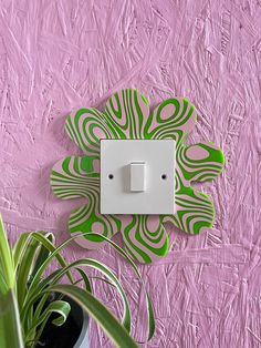 a green and white flower shaped light switch plate on a pink wall next to a potted plant