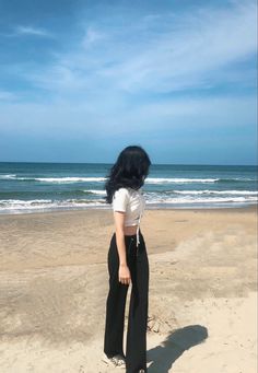 a woman standing on top of a sandy beach next to the ocean with her hands in her pockets