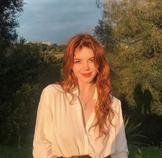 a woman with long red hair wearing a white shirt and brown belt standing in front of trees