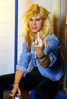 a woman with blonde hair sitting on a stool pointing at the camera and making a peace sign