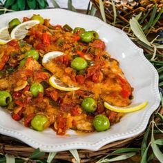 a white plate topped with fish covered in sauce and olives next to lemon wedges