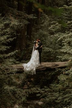 a bride and groom standing on a fallen log in the woods, hugging each other