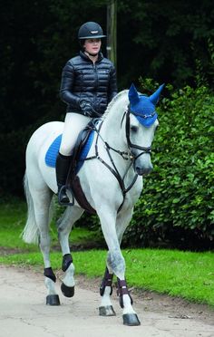 a woman riding on the back of a white horse down a dirt road next to trees