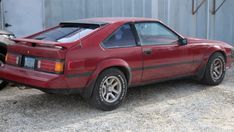 a red sports car parked in front of a metal building next to two cars on gravel