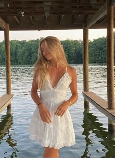 a woman in a white dress is standing on a dock by the water with her hands behind her back