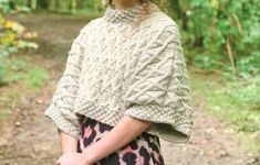 a woman standing in the woods wearing a knitted sweater over her shoulders and looking off into the distance