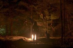 people standing around a campfire in the dark forest at night with one person jumping up into the air