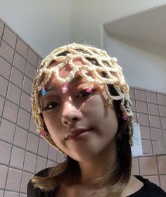 a woman wearing a crocheted hat with beads on her head and eyes, in front of a tiled wall