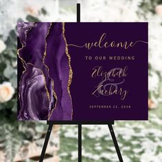 a purple and gold wedding welcome sign on an easel with flowers in the background