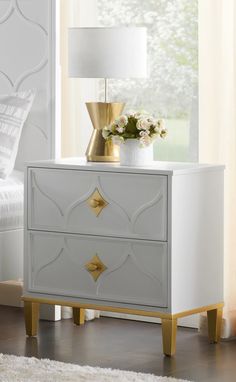 a white and gold nightstand with flowers on it in front of a window, next to a bed