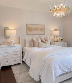 a white bed sitting under a chandelier next to two tables with lamps on it