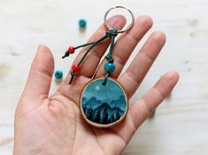 a hand holding a wooden keychain with mountains in the background and beads around it