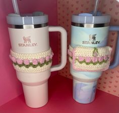 two coffee mugs with crocheted sleeves and matching cups in front of a pink background