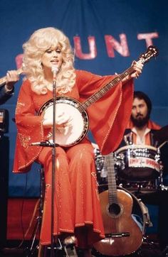 a woman with blonde hair playing a musical instrument and singing into a microphone while another man plays the guitar behind her