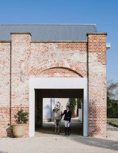 a woman standing in front of a brick building with an open door and a horse