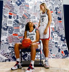 two female basketball players sitting on chairs in front of a wall with images of women's basketball teams