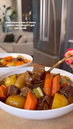 two white bowls filled with stew and carrots on top of a wooden table next to a woman's hand