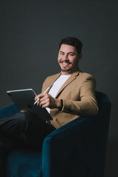 a man sitting in a blue chair holding a laptop computer and smiling at the camera