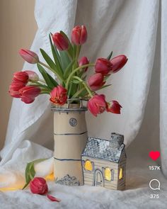 red tulips are in a vase next to a small house on a white cloth