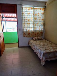 a small room with a bed, window and door leading to another room that has tile flooring