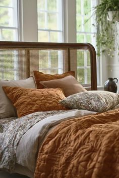 a bed with pillows and blankets in front of two large windows that have potted plants on them