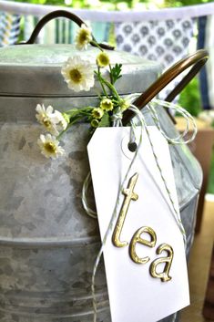 a tea kettle with a tag hanging from it's handle and some flowers in the background